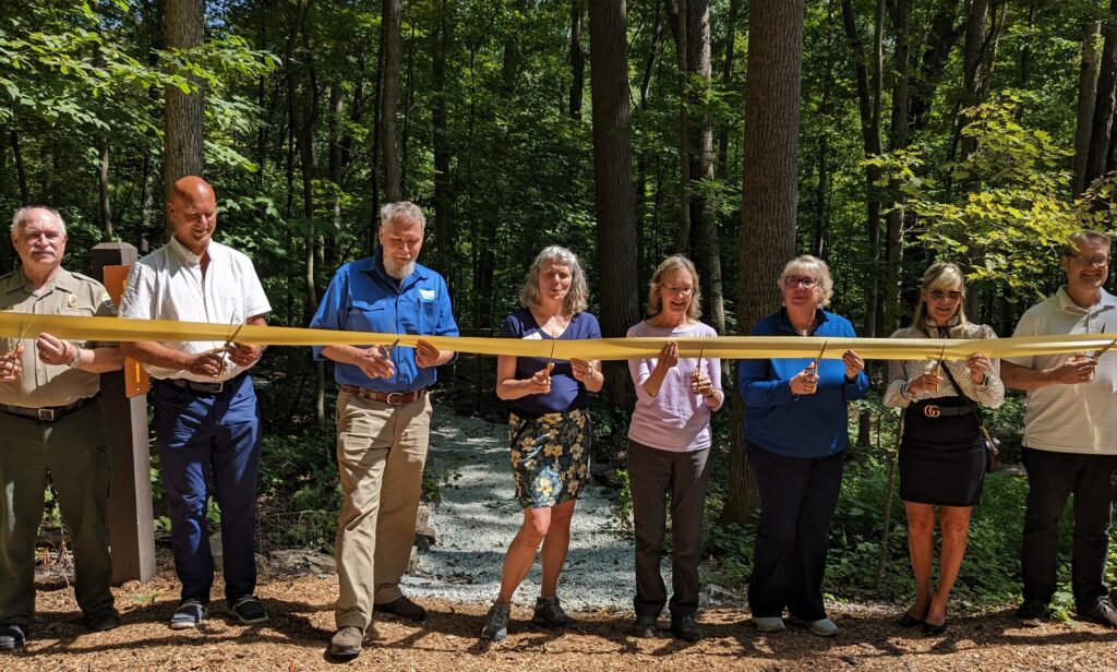 Pictured is 8 people holding a ribbon and scissors. They are standing in front of a stand of trees on a mulched path with a new gravel path that leads to the new campsite behind them.