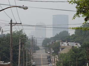 Photo shows a city scape in Pittsburgh from a road on a hill. The buildings are barely visible though thick smoke from the summer 2023 Canadian Wildfires. 