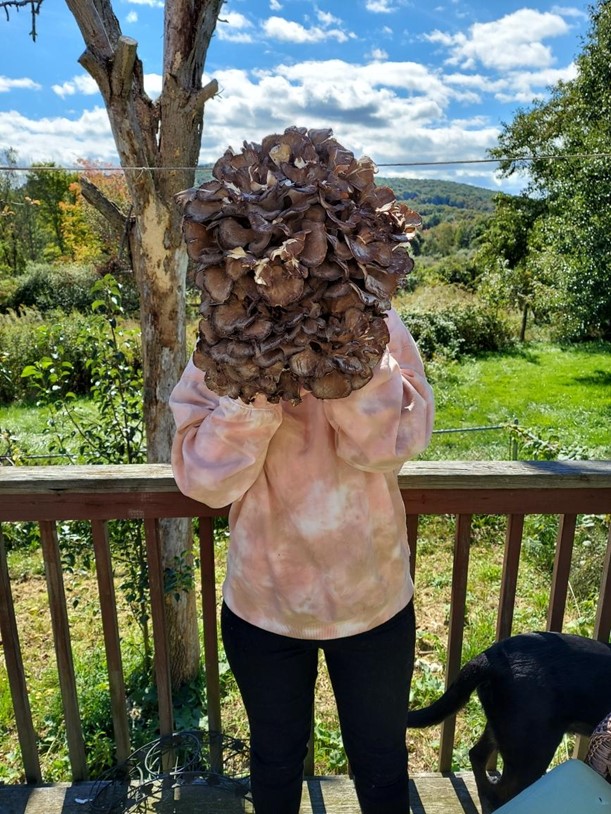 A cluster of mushroom "petals" are being held up in front of a woman and it is twice the size of her head. She is wearing a pink and white marbled sweatshirt and black leggings. She is leaning against a porch rail and a black dog stands next to her. There is a yard full of vegetation in the background that fades into a mountain background with blue sky and fluffy white clouds. 
