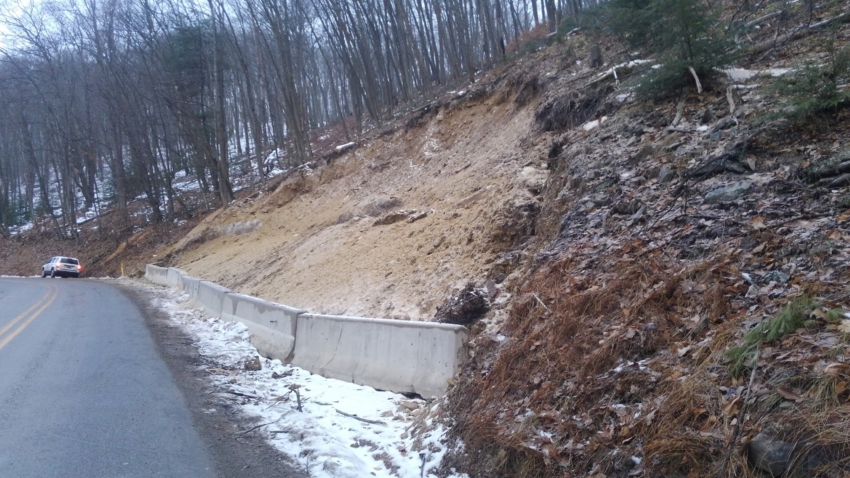 Image shows a cement wall barrier along a road to block any further mudslide from blocking the road. It is a side hill of dirt and snow and trees bare from the winter