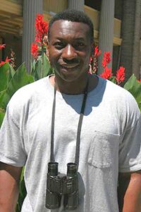 Keith Russell posing with his binoculars. He is a black man in a grey tshirt. Behind him is a closely copped garden showing a few red flowers. 