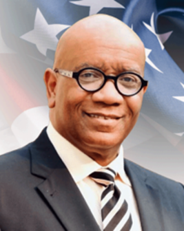 Brian K Wade in a black man in a black suit, white shirt, and black and white diagonally slanting lines. He wears back framed glasses and is bald. The background is an image of the American Flag