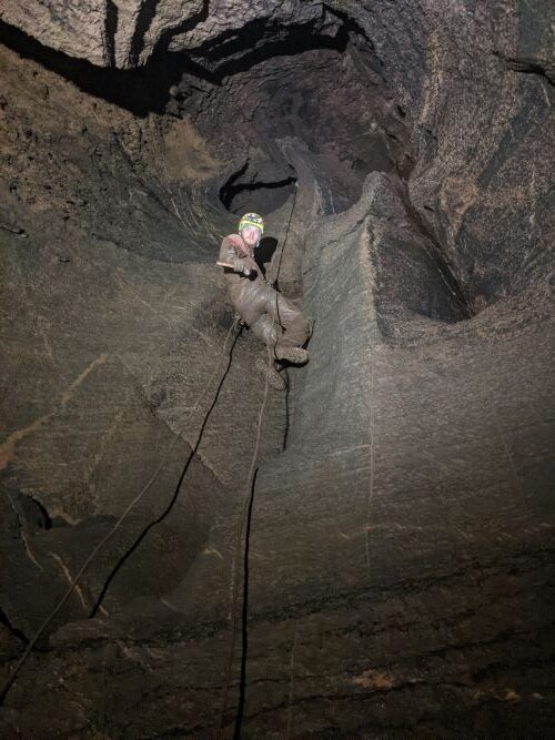 A person covered in mud hangs from the small opening on a climbing rope of a hole in the top of a cave. 