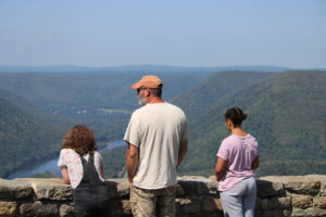 Two preteen girls and a middle aged man look out across a gorge and river from an overlook. The girl on the left has shoulder length brown curly hair and is in a floral teeshirt and dark bib overalls. The man in the middle has an orange ball cap and salt and pepper hair and beard in a white tshirt and khaki camo pants. The girl on the right has dark wavy hair pulled into two buns and is wearing a lilac colored tshirt and medium blue pants. The girl on the left and man in the middle are white with tans and the girl on the right in a light to medium brown complexion. 